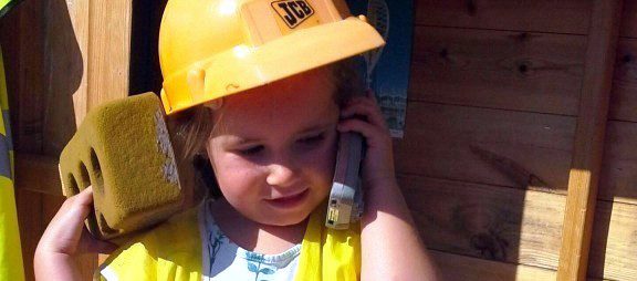 toddler girl wearing a dress-up construction helmet and high vis holding a toy phone to her ear